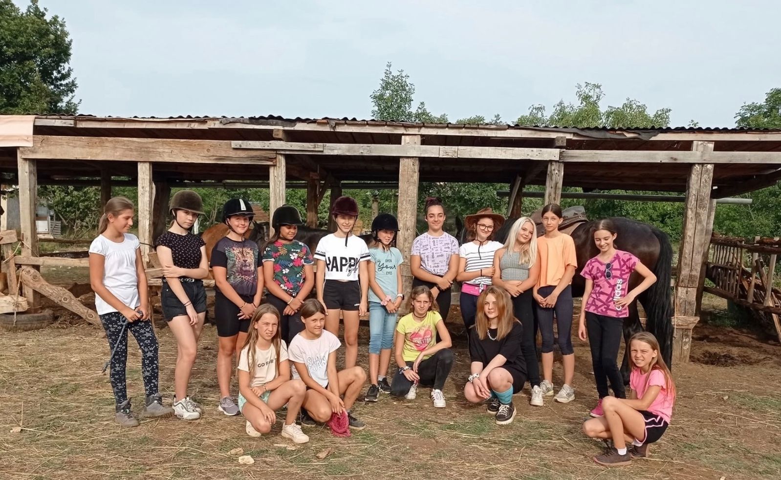 A group of young riders gathered in front of the wooden stable shelter, with the horses behind them