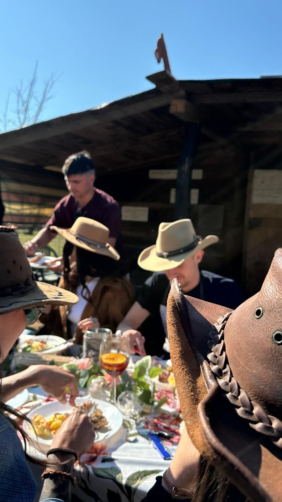 Guests sharing a meal at a long outdoor table beside the wooden stable on a sunny day