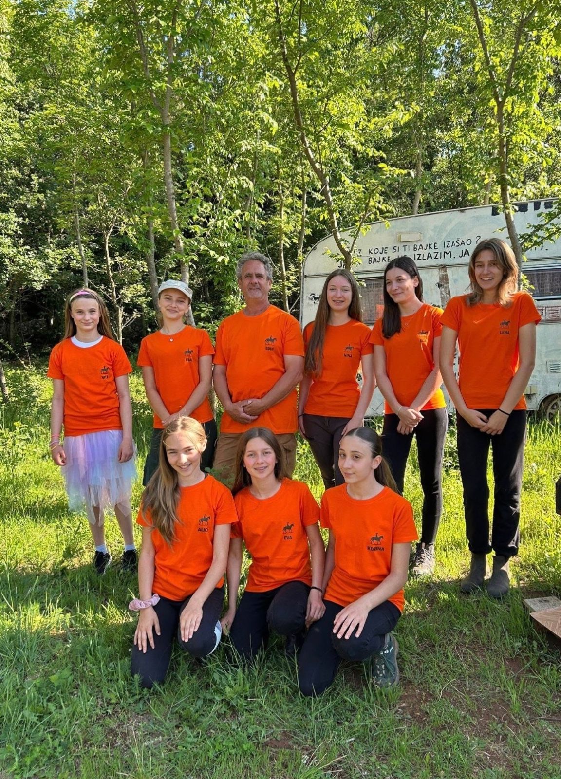 The riding school group in matching orange ranch t-shirts, standing together in the grass under the trees