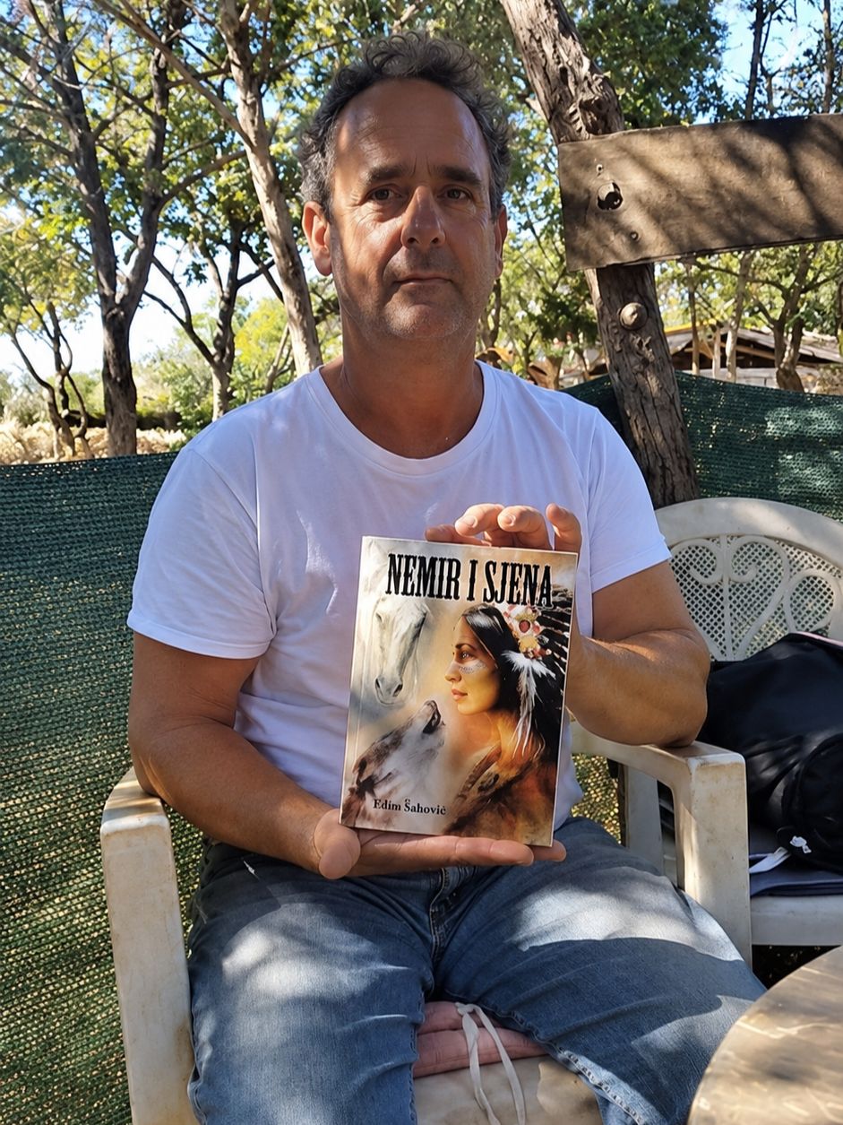 The founder sitting at the ranch, holding a copy of his novel Nemir i Sjena