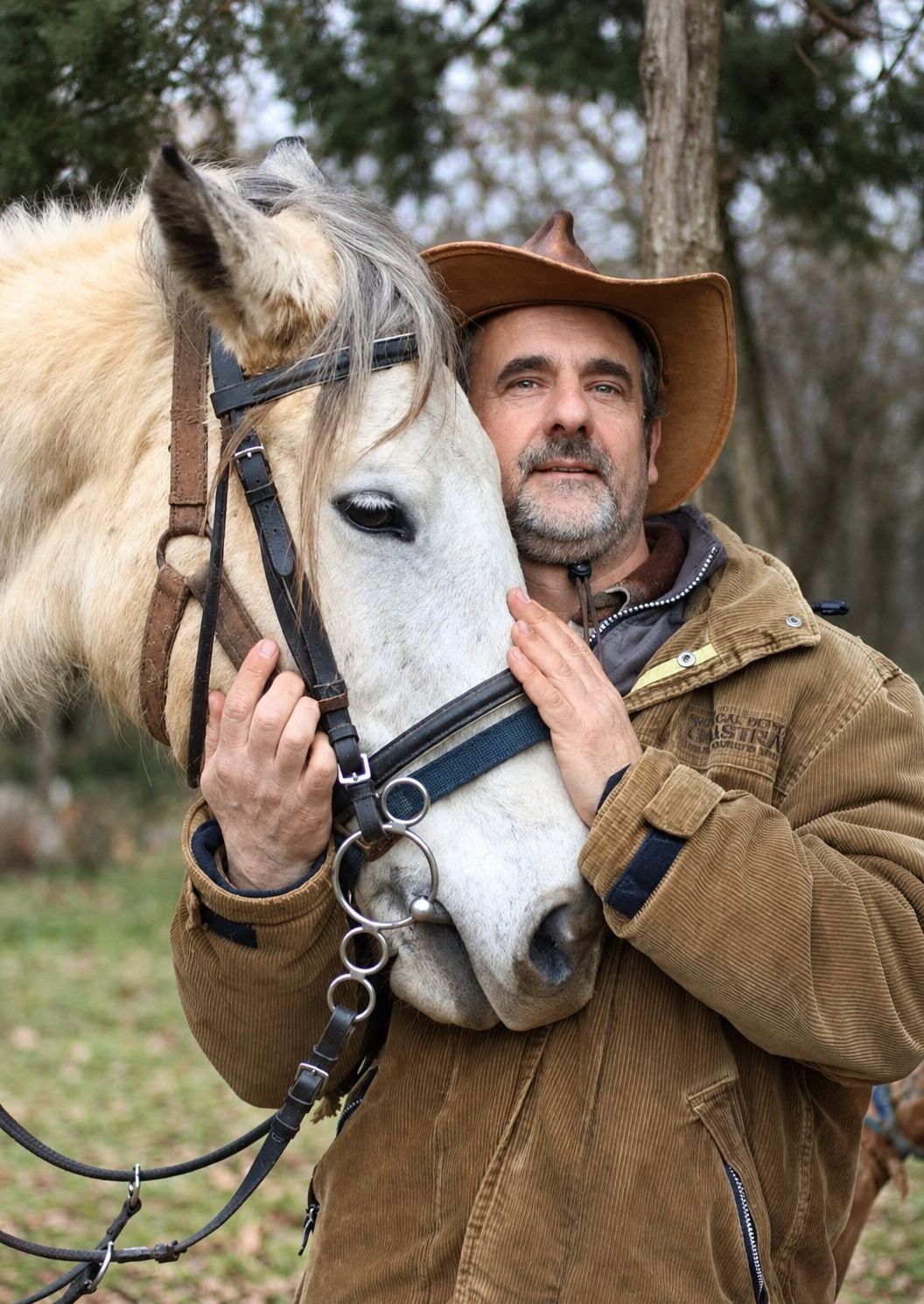 The founder of the ranch holding the head of a pale horse close to his face, among the trees